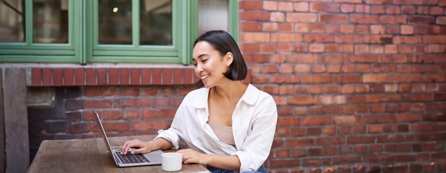 Stylish young businesswoman, asian girl with laptop, sitting in outdoor cafe with cup of coffee and working, using computer.