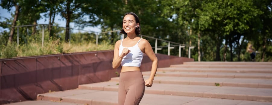 Wellbeing and sport. Asian fitness girl runner, jogging in park, running on street in leggings, smiling happily.