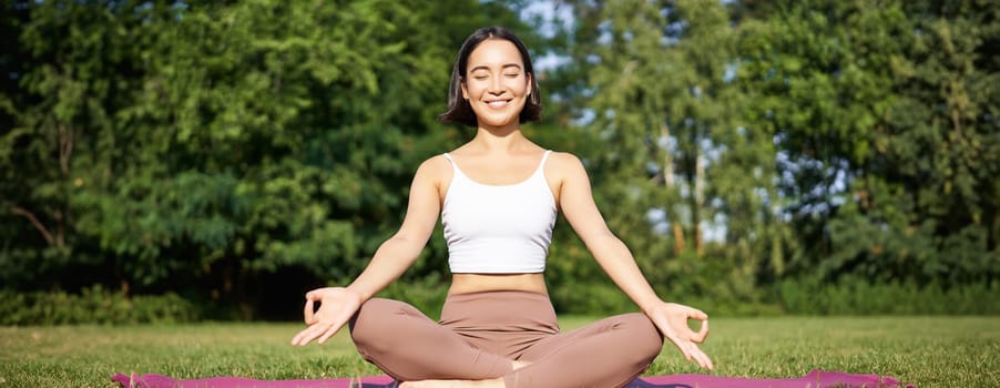 Sporty woman meditating on fresh air, sitting on fitness mat and practice yoga, smiling pleased. Sport and people concept.