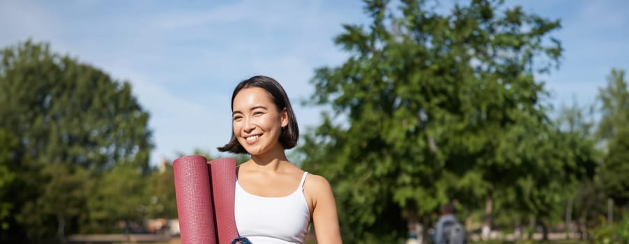 Portrait of young slim and healthy korean girl doing workout in park, standing with water bottle and rubber mat for execises on green lawn, smiling happily.