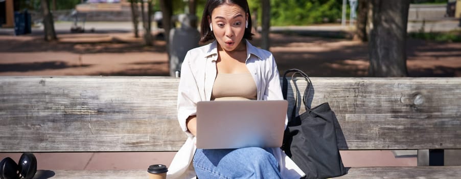 Happy asian girl chatting, having video call on laptop, sitting on bench in park and communicating, talking to someone online.