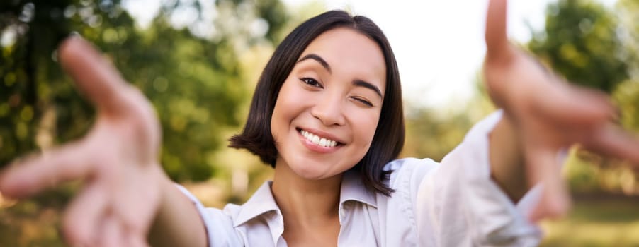 People and mobile connection. Happy young asian woman takes selfie on smartphone, holds camera with hands, poses in park on summer day.