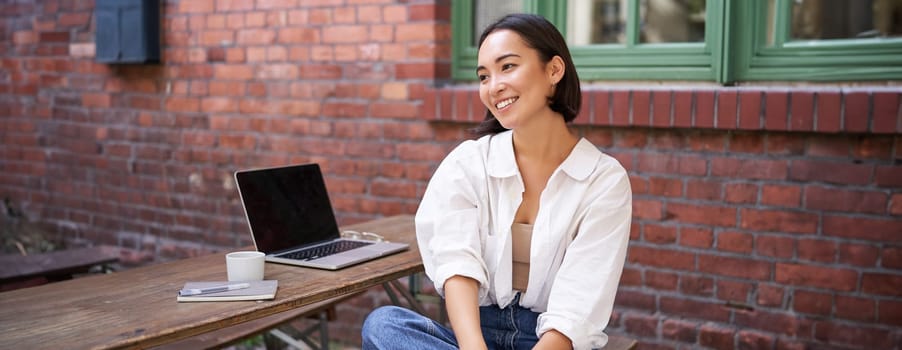 Relaxed and happy asian woman, sitting with laptop in outdoor cafe, drinking coffee, smiling and laughing.