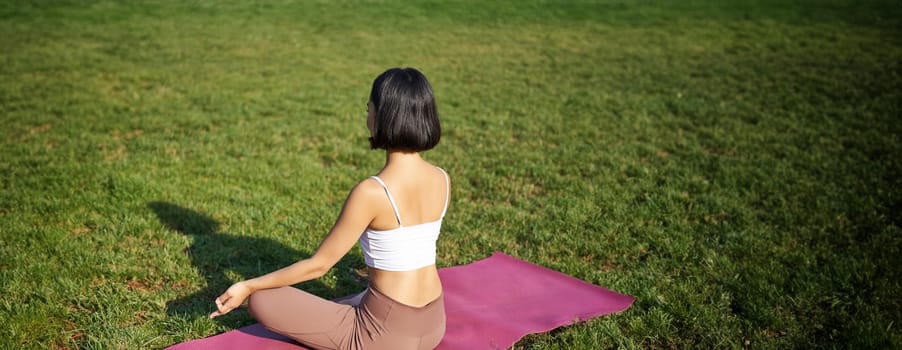 Rear view of woman silhouette doing yoga, sitting on fitness mat and meditating on green lawn.