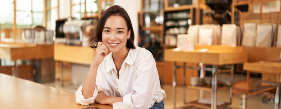 Portrait of confident asian woman, sitting in cafe, smartphone and coffee on table. Businesswoman smiling with confidence. Copy space