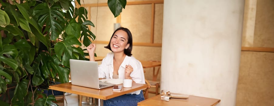 Happy young asian woman winning on laptop, receive good news, achieve goal at work, triumphing and smiling pleased, sitting in cafe.
