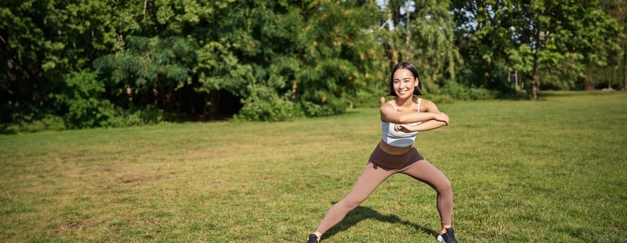 Young asian fitness girl stretching, workout in park, showing exercises, doing squats.
