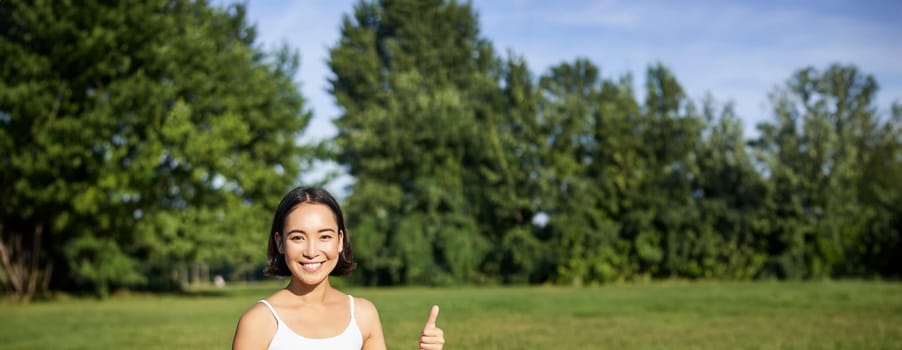 Vertical shot of asian woman shows thumbs up, recommending yoga training online, meditation app, doing exercises on fresh air in park.