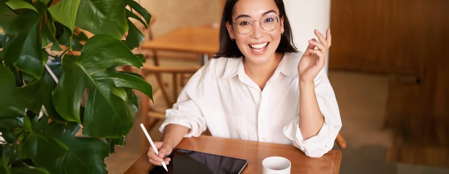 Young asian creative girl, sitting with digital tablet, drawing with graphic pen and smiling, doodling, drinking coffee in cafe.