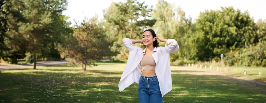 Carefree asian girl laughing and dancing in park, enjoying summer sunny day, raising hands up and breathing fresh air.