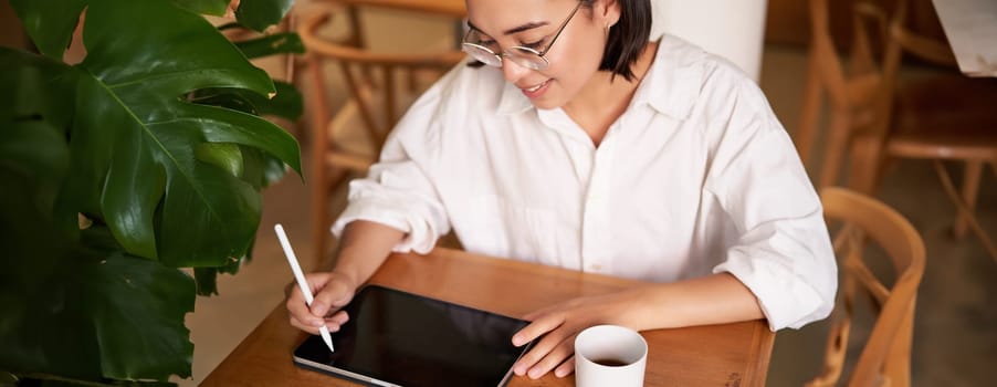 Young asian creative girl, sitting with digital tablet, drawing with graphic pen and smiling, doodling, drinking coffee in cafe.