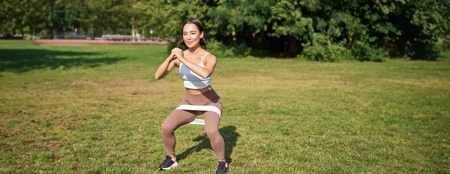 Woman using stretching band, resistance fitness rope for workout in park, doing squats, training on fresh air.