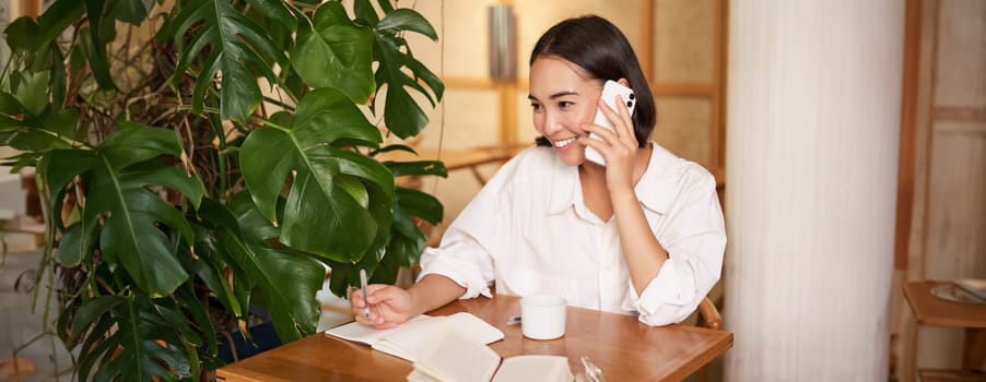 Working woman answer phone call in cafe, writing down, making notes while having conversation on telephone.