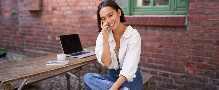 Beautiful korean girl with laptop, sitting in outdoor cafe, drinking coffee and using computer. Young people and recruitment concept