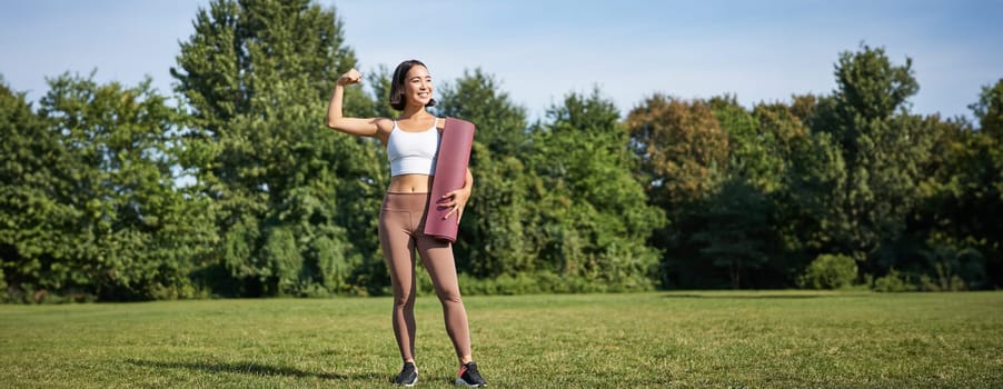 Portrait of fit and healthy young fitness woman, shows her biceps with pleased smiling face, stands with yoga rubber mat on grass in park.