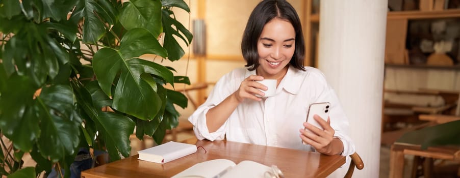 Young asian businesswoman sitting in cafe with notebook, coffee and looking at smartphone, video chat, calling someone.