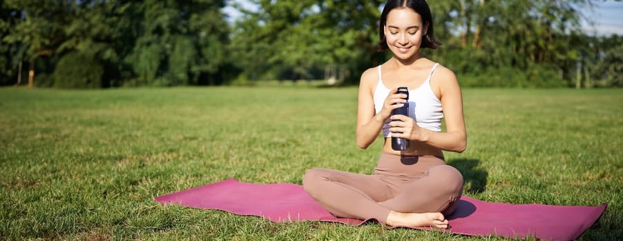 Smiling fitness girl sits on mat and drinks water after workout, finish yoga training in park, exercise on fresh air.