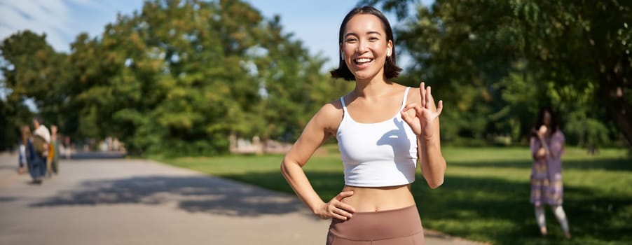 Smiling korean girl jogger, shows okay, ok sign, approval gesture, say yes, standing in park during workout.