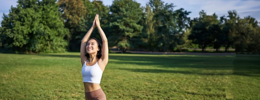 Vertical shot of young asian fitness girl doing yoga asana, standing in tree pose and smiling, execising in park on rubber mat.