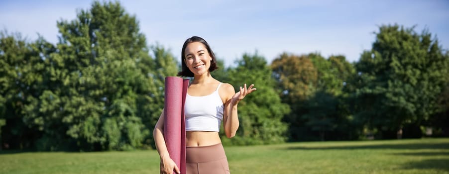 Excited asian girl in sportswear, holds rubber mat for yoga, looks surprised and happy, stands in park, workout on fresh air outdoors.