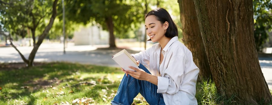 Beautiful young asian girl, student sits in park under tree and reading book, smiling, enjoying warm summer day outdoors.