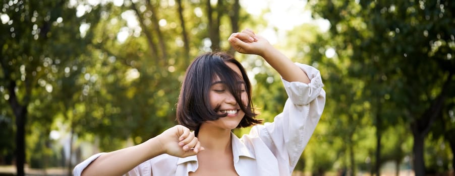 People lifestyle. Portrait of young brunette woman dancing, smiling and laughing, walking in park with hands lift up high, enjoying summer day outside.