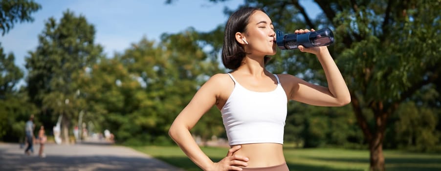 Smiling asian fitness girl drinks water, workout in park, stay hydrated during jogging training session.
