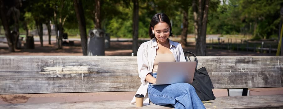 Portrait of korean woman sitting with laptop in park on bench, working outdoors, student doing homework, drinking takeaway coffee.