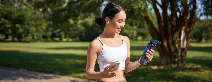 Portrait of fitness girl, runner drinking water, looking at smartphone, standing in park in middle of workout, sport training session.