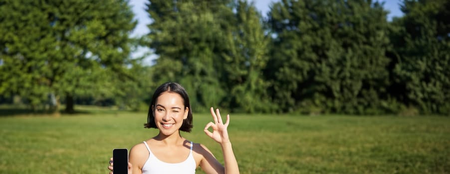 Vertical shot of asian woman shows okay sign and smartphone screen, recommending yoga training online, meditation app, doing exercises on fresh air in park.