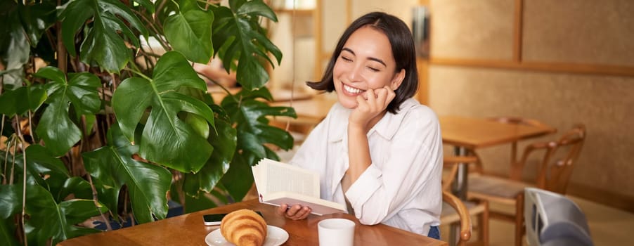 Romantic asian woman sitting with book in cafe, eating croissant and drinking coffee, reading and smiling, enjoying alone time.