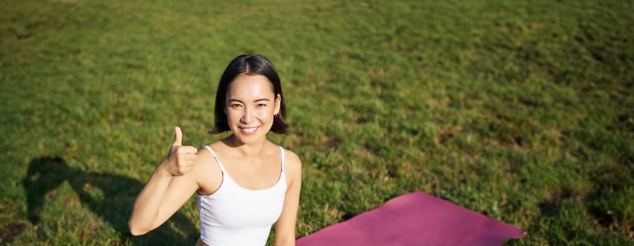 Vertical shot of asian woman sitting on rubber mat after good yoga training in park, showing thumb up in approval. Meditating on fresh air.