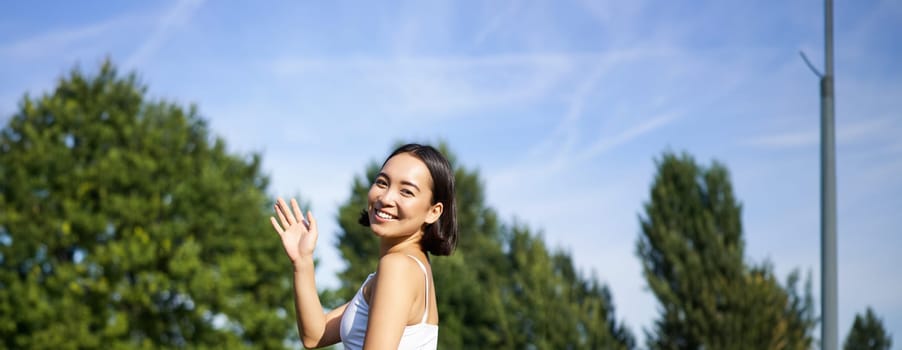 Portrait of asian girl on fitness class in park, sitting on rubber mat and wave hand at camera, say hello, meditating and practice yoga.