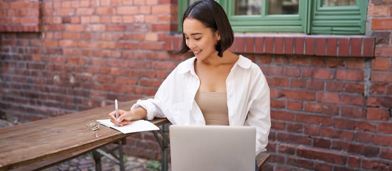 Working woman sitting outdoors in cafe, looking at laptop and making notes in notebook, writing down while watching smth on computer.