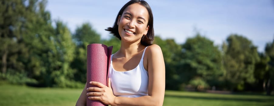 Portrait of asian girl smiles and laughs, stands with sports equipment, rubber mat, wears uniform for workout and exercises in park.