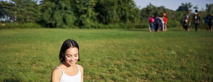 Portrait of asian girl sitting on mat in park, talking to smartphone, meditating online with yoga instructor on fresh air.