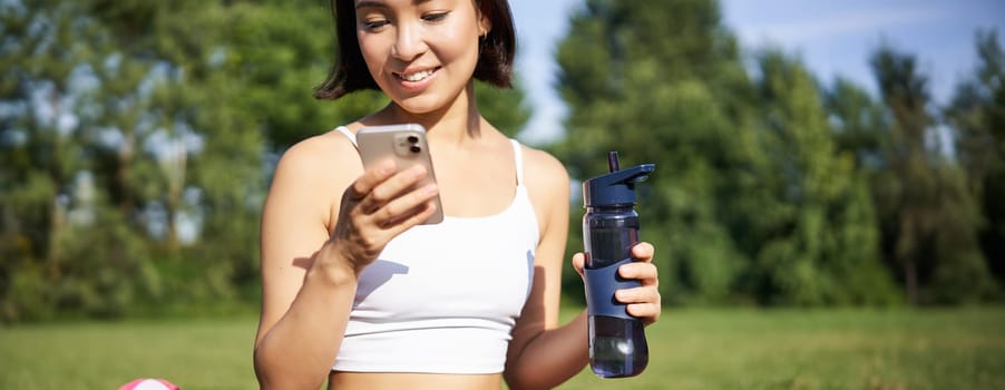 Smiling fitness girl drinks water, checks her app on smartphone and looks happy, stays hydrated on fresh air, sunny day in park.