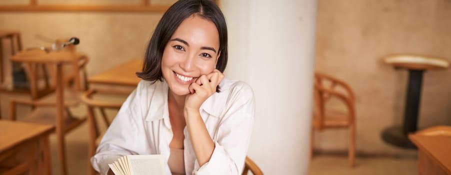 Vertical shot of happy young asian woman enjoys reading, sitting with book in cafe, drinking coffee and eating croissant.