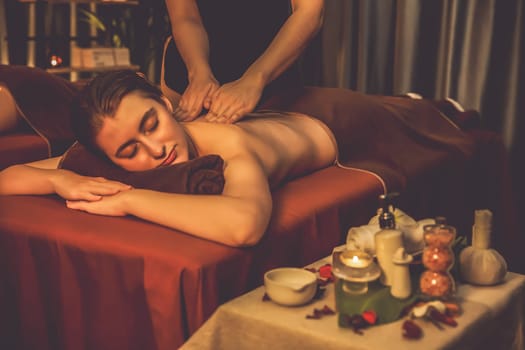 Caucasian couple customer enjoying relaxing anti-stress spa massage and pampering with beauty skin recreation leisure in warm candle lighting ambient salon spa at luxury resort or hotel. Quiescent