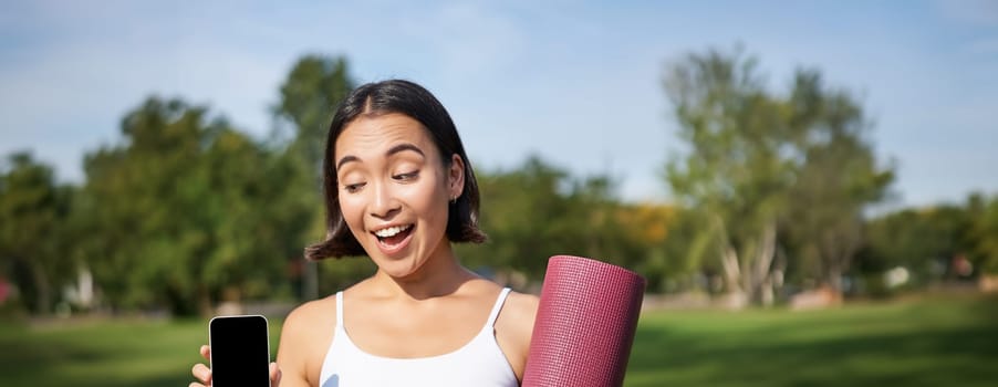 Excited fitness girl recommends application for sport and workout, shows phone screen, standing with rubber yoga mat in park after training session.