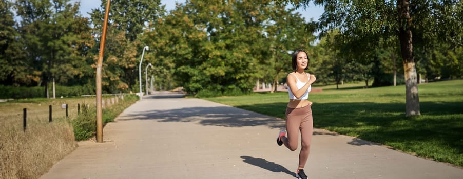 Young asian fitness woman running in park, jogging outdoors, smiling pleased.