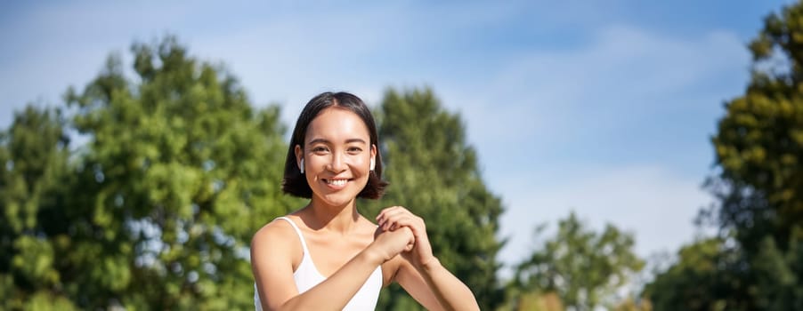 Asian fitness girl doing squats in park, using resistance band, stretching yoga rope for workout training on fresh air.