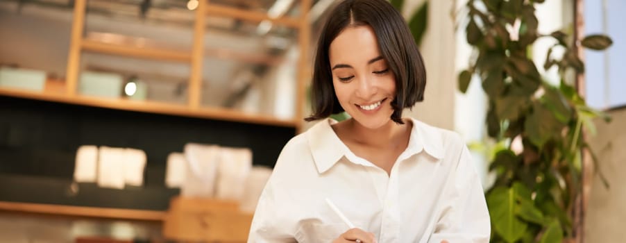 Portrait of asian girl drawing on digital tablet, scatching and doodling with graphic pen, sitting in cafe and smiling.