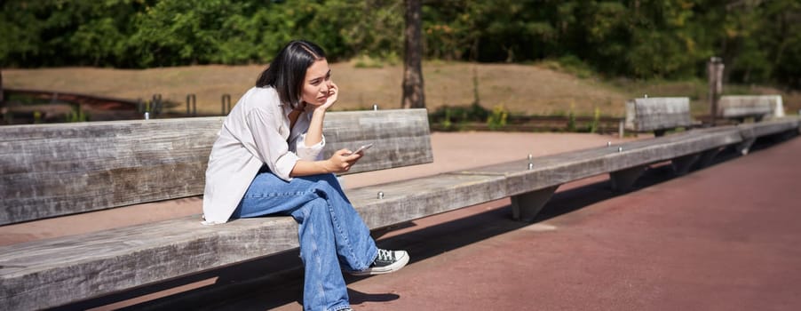 Portrait of asian girl sitting with smartphone feeling sad, looking gloomy and frustrated, waiting for a call outdoors in park.