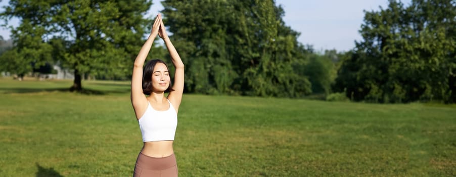 Smiling young fitness girl on rubber mat, workout in park, raising hands up in tree pose, doing yoga training on fresh green lawn.