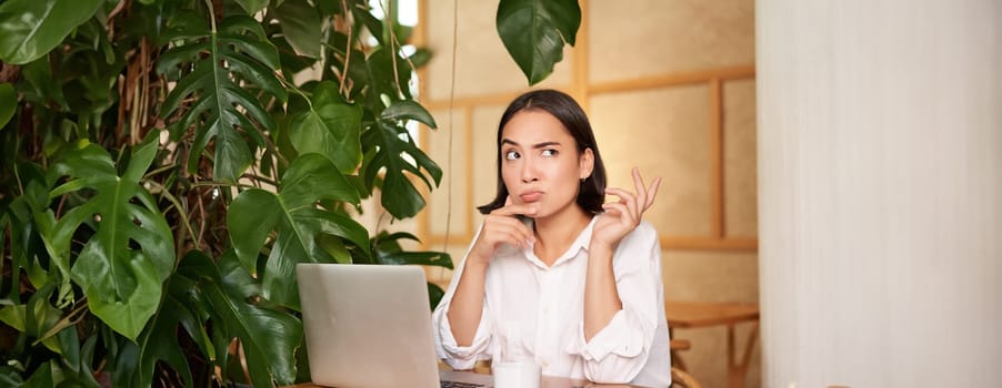 Young businesswoman thinking, frowning as sitting puzzled and complicated, using laptop in cafe.