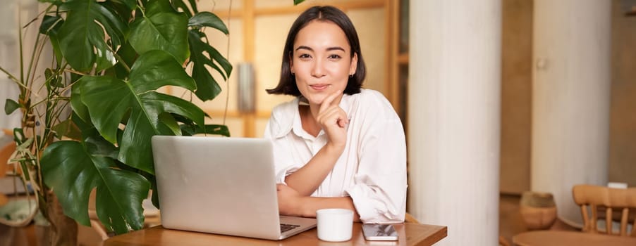 Confident young stylish woman with laptop, sitting in cafe and working, freelancer in co-working space with cup of coffee.