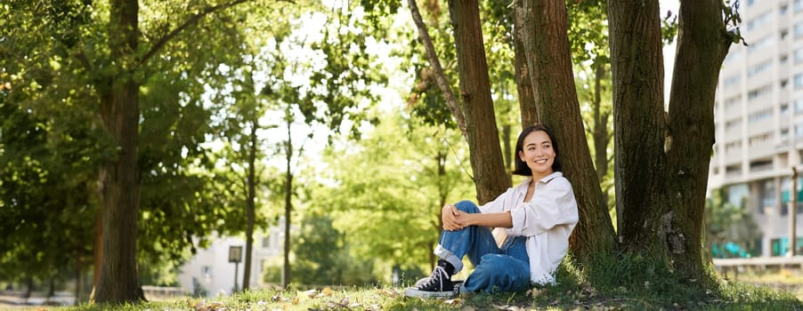 Beautiful smiling girl sits near tree in park, enjoying nature outdoors, relaxing and resting on fresh air. Copy space