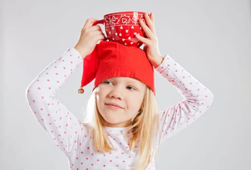 Studio portrait of smiling young girl wearing red Christmas elf hat holding big red cup on head