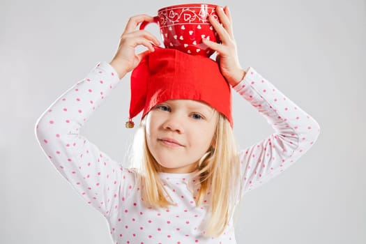 Studio portrait of smiling young girl wearing red Christmas elf hat holding big red cup on head